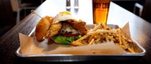 A burger with fried pickles and an egg, french fries, and a glass of beer with the tipsy cow logo on it